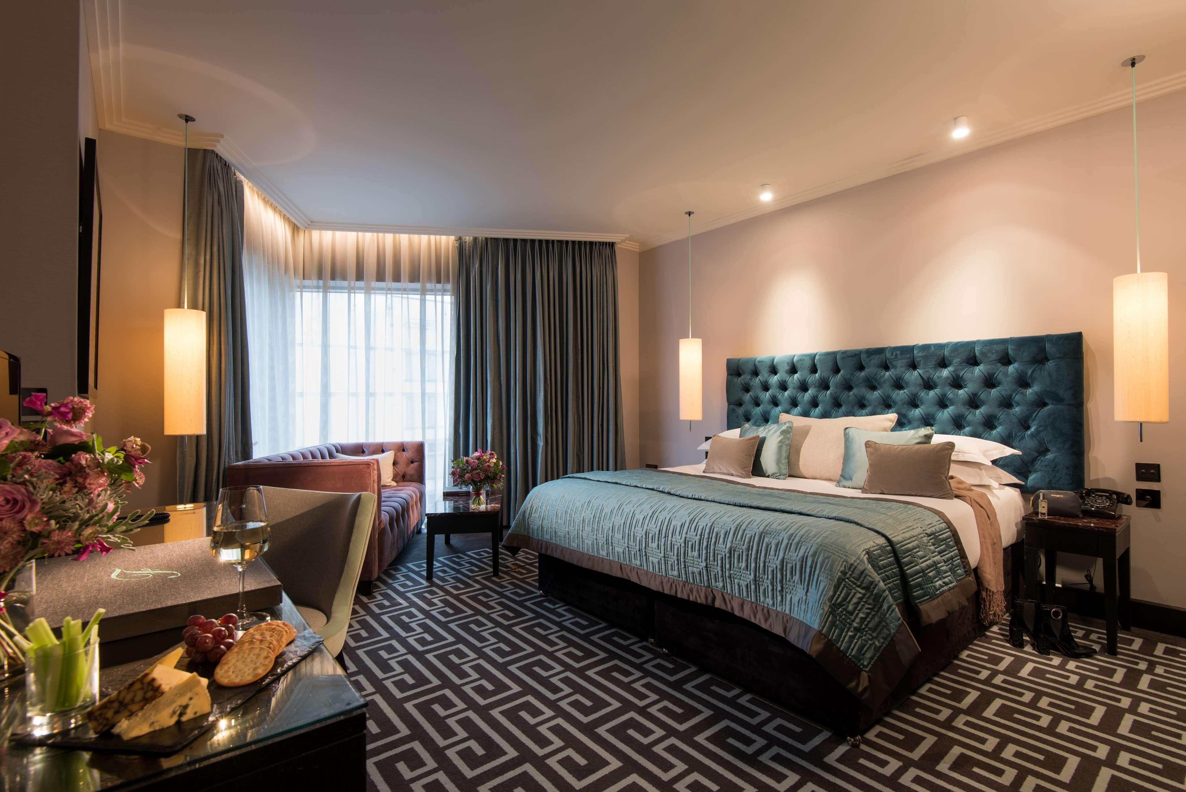 The Fitzwilliam Hotel Belfast Bagian luar foto The photo depicts a stylish hotel room. It features a large bed with a tufted teal headboard, flanked by two wall sconces providing soft lighting. The bedding is neatly arranged with accent pillows. To one side, there is a small seating area with a c