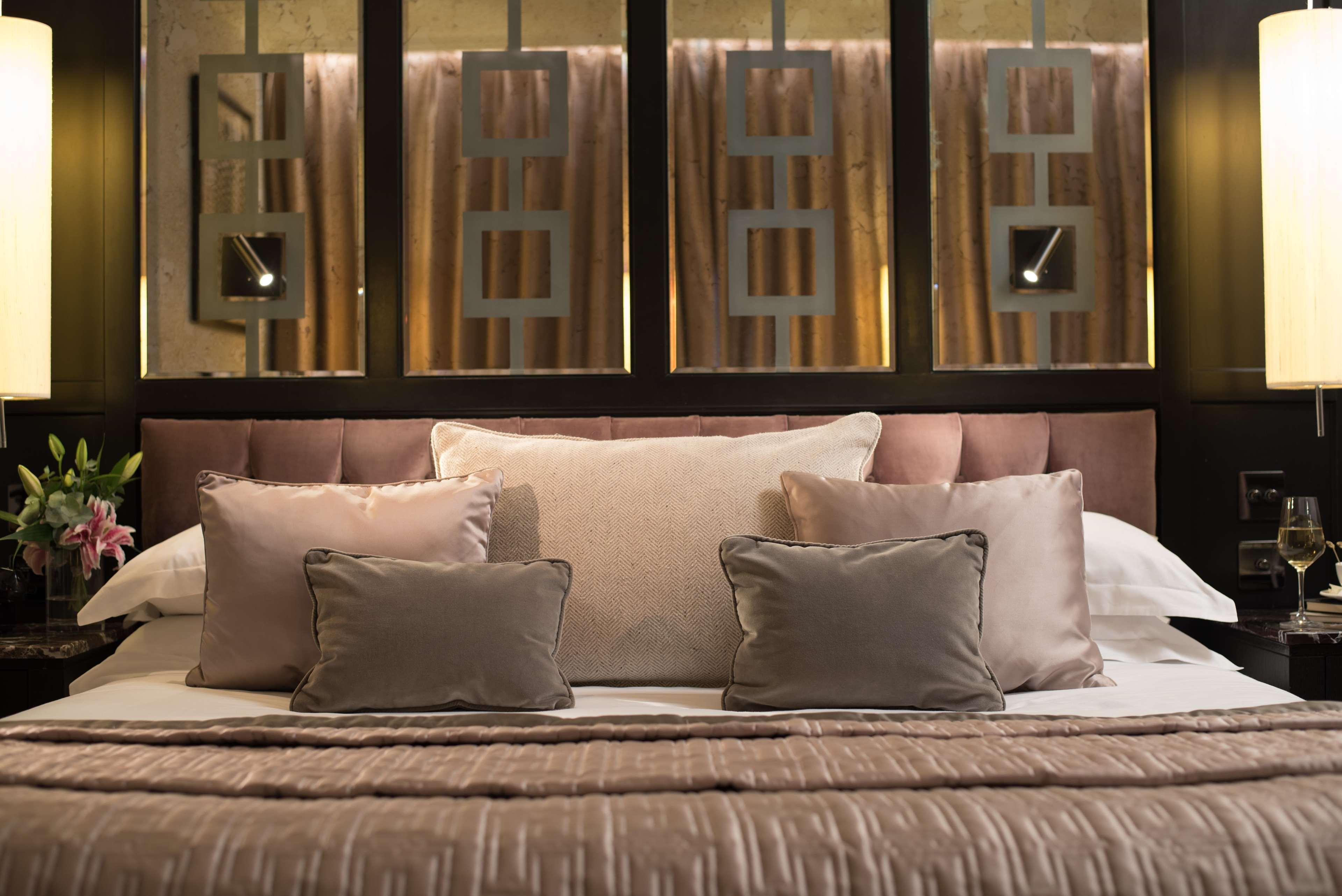 The Fitzwilliam Hotel Belfast Bagian luar foto The photo shows a beautifully designed bed in a stylish bedroom. The bed is adorned with a soft, plush headboard and layered with various decorative pillows in shades of beige and gray. The bedding appears luxurious, with a textured blanket. Behind t