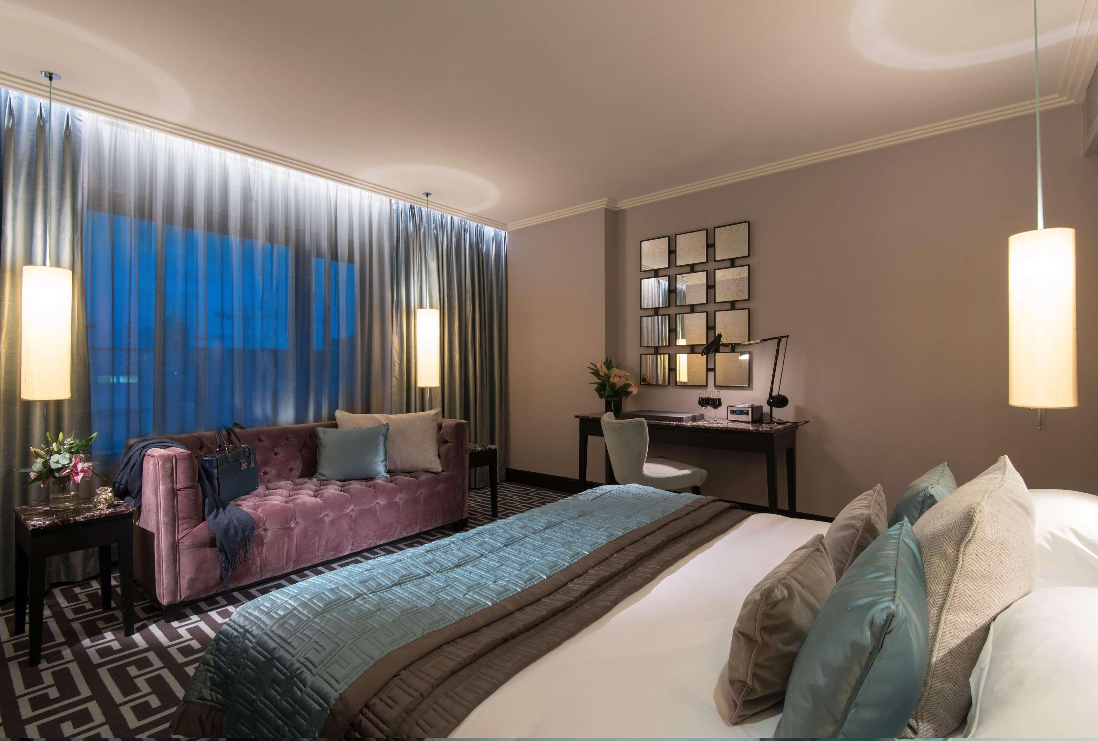 The Fitzwilliam Hotel Belfast Bagian luar foto The photo depicts a modern hotel room featuring a spacious layout. There is a large bed with decorative pillows in the foreground, covered with a stylish bedspread. To one side, a plush purple sofa adds a pop of color and comfort. A small desk and ch